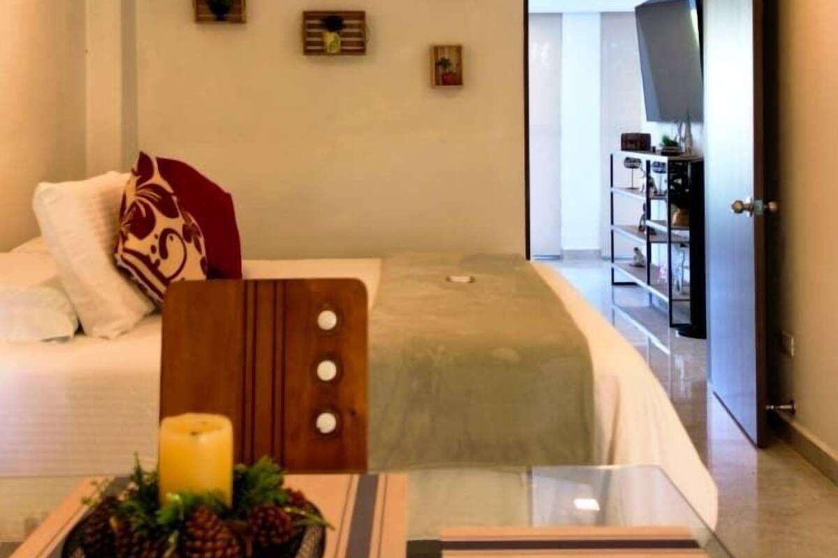 A modern hotel room with a bed, a small table with a candle and pine cones, a sofa, and a view into the hallway.