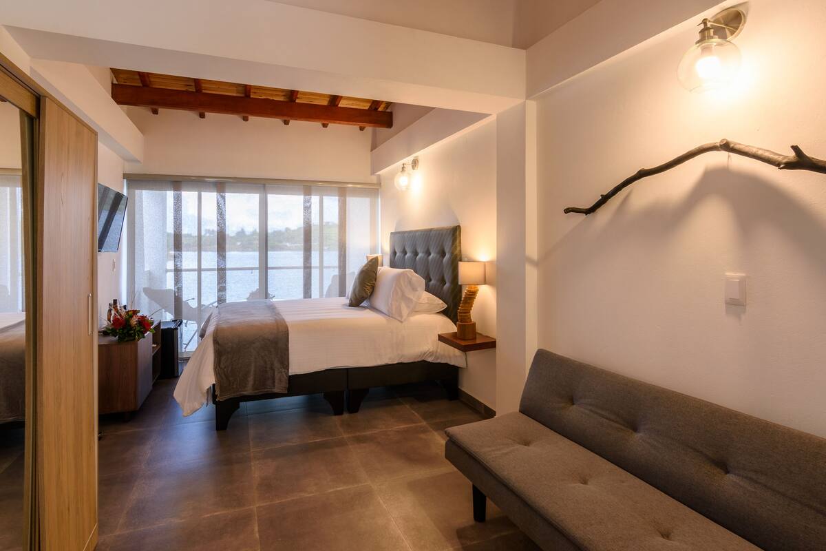 A bedroom with a large bed, a sofa, a desk with a chair, and a window with a view.