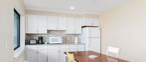Una cocina compacta con armarios blancos, microondas, refrigerador y una mesa de comedor con una silla.