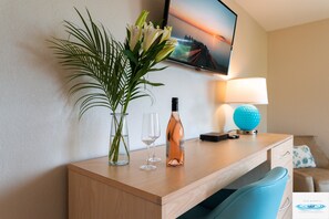 A hotel room with a wooden desk, a TV mounted on the wall, a vase with flowers, two wine glasses, and a bottle of wine.