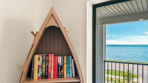 A triangular wooden bookshelf filled with books, positioned next to a door with a view of the sea.