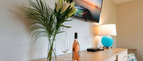 A hotel room with a wooden desk, a vase with flowers, a wine bottle, two glasses, a television, a lamp, and a blue decorative object.