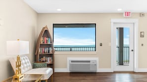 A hotel lobby with a large window overlooking a body of water, a bookshelf, a seating area with a lamp, and an air conditioning unit. - 🏖️ Gold Coast Inn | Breathtaking Lake Views • Corner Rm 201 (Traverse City)