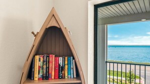 A triangular wooden bookshelf filled with books, positioned next to a door with a view of the sea. - 🏖️ Gold Coast Inn | Breathtaking Sunsets w/ Lake View ➠ 106 (Traverse City)