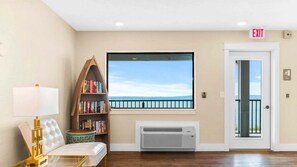 A room with a large window, a bookshelf, a white door with an exit sign, and a small table with a lamp.