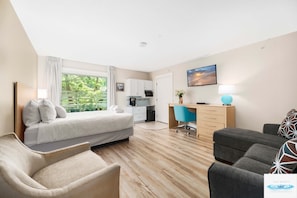Habitación de hotel con cama, escritorio, silla, sofá, kitchenette pequeña y ventana con cortinas.