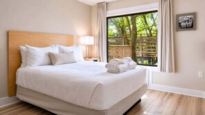 Une chambre d’hôtel avec un grand lit, une table de chevet, une lampe, une fenêtre avec des rideaux et une vue sur les arbres.