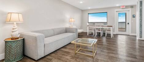 Una sala de estar moderna con un sofá, una mesa de centro y lámparas. Hay un comedor con sillas y una mesa, y una puerta que da al exterior.