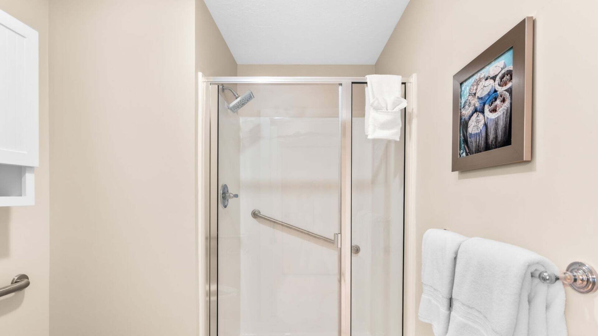 A glass-enclosed shower with a handrail, a white towel hanging on the wall, and a framed picture above the towel.