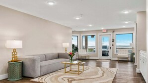 A hotel lobby with a grey sofa, two lamps, a glass coffee table, and a patterned rug. - 🏖️ Gold Coast Inn | Breathtaking Sunsets w/ Lake View ➠ 303 (Traverse City)