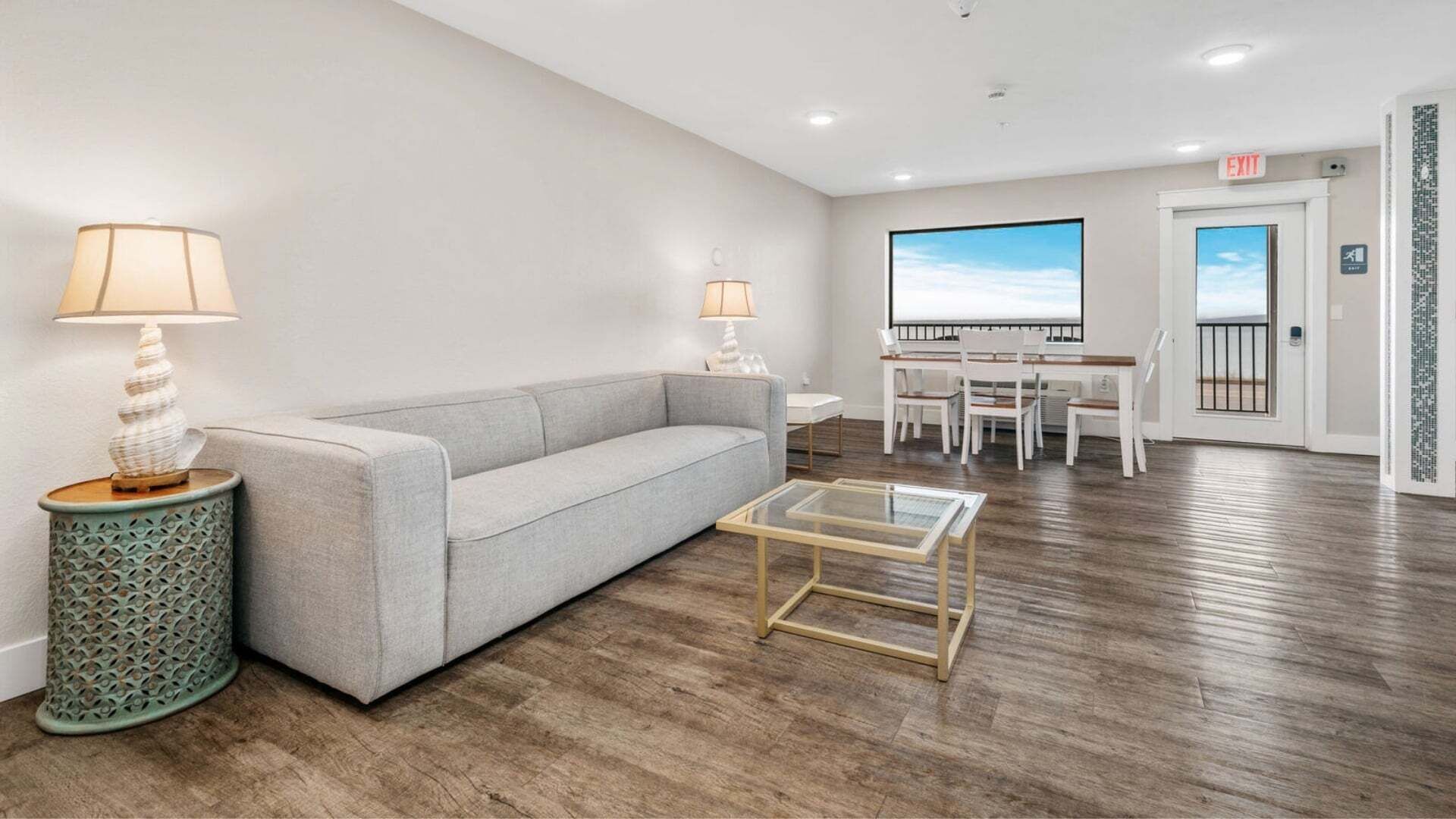 Uma sala de estar moderna com sofá, mesa de centro e luminárias. Há também uma área de jantar com cadeiras e uma mesa, e uma porta que dá para o exterior.