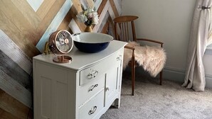 A corner room with a herringbone-patterned wall, a white dresser with drawers, a blue bowl on top, a vintage fan, a chair with a fluffy seat, and a small plant hanging on the wall. - Nemo Farmhouse on Centennial Trail With a View (Nemo)