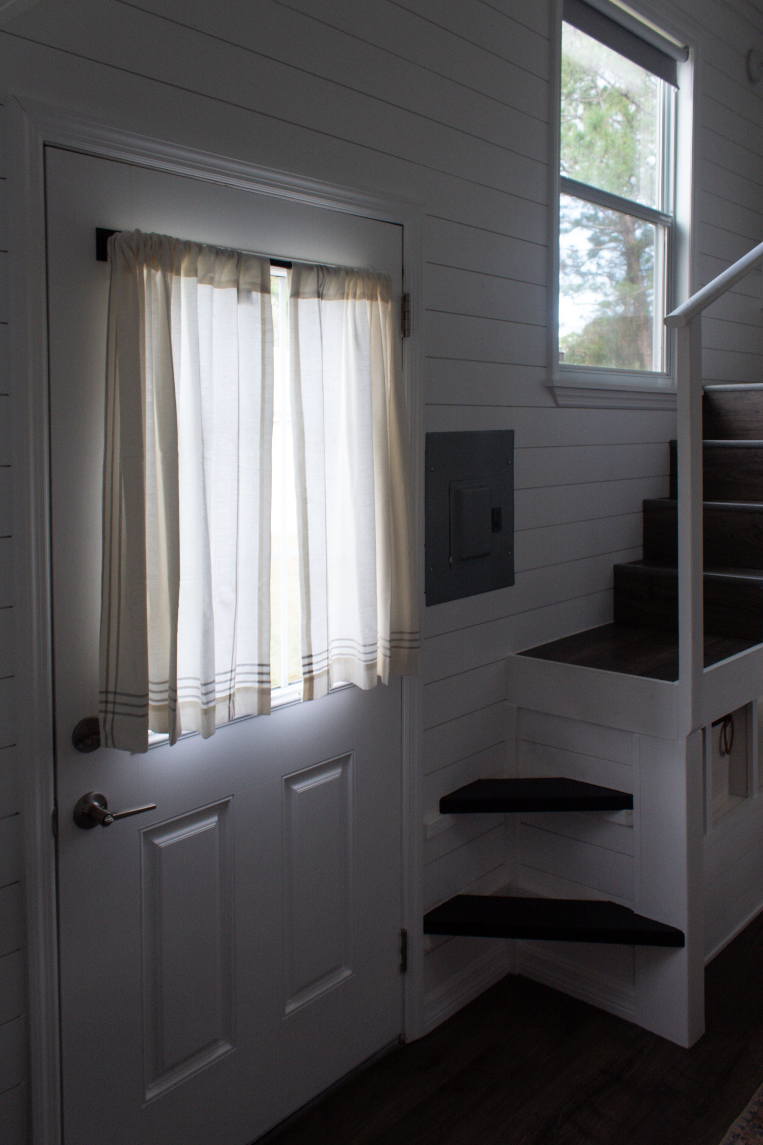 A white door with a window above it, a small window with a view of trees, and a dark wooden staircase with a handrail.
