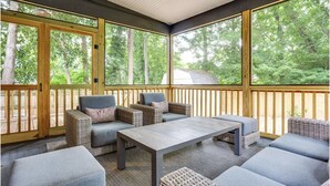 Un patio cubierto con mobiliario de mimbre, una mesa y ventanas amplias que dan a los árboles circundantes.