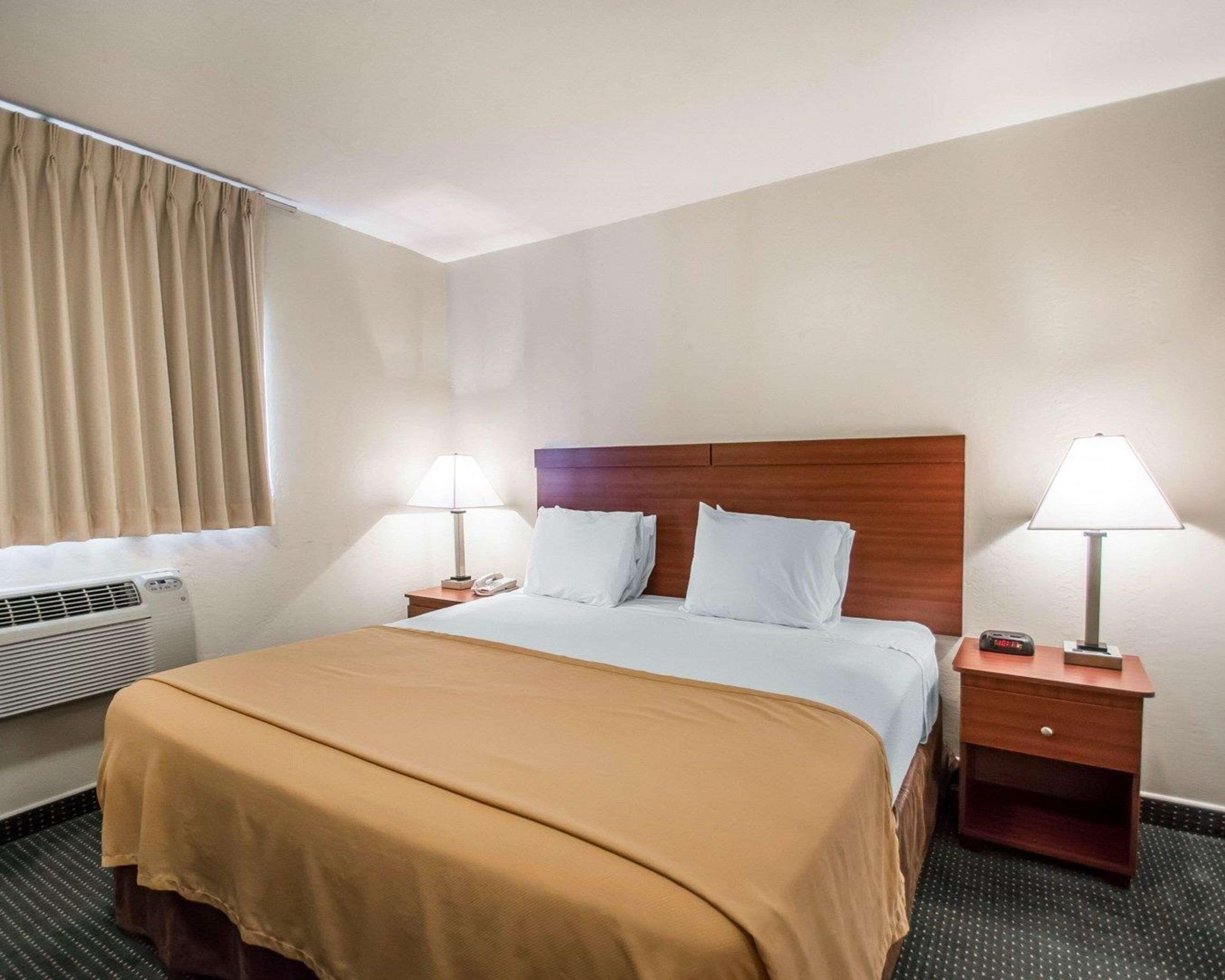 Habitación de hotel con una cama, dos mesitas de noche, una lámpara sobre cada una, una ventana con cortinas y un equipo de aire acondicionado.