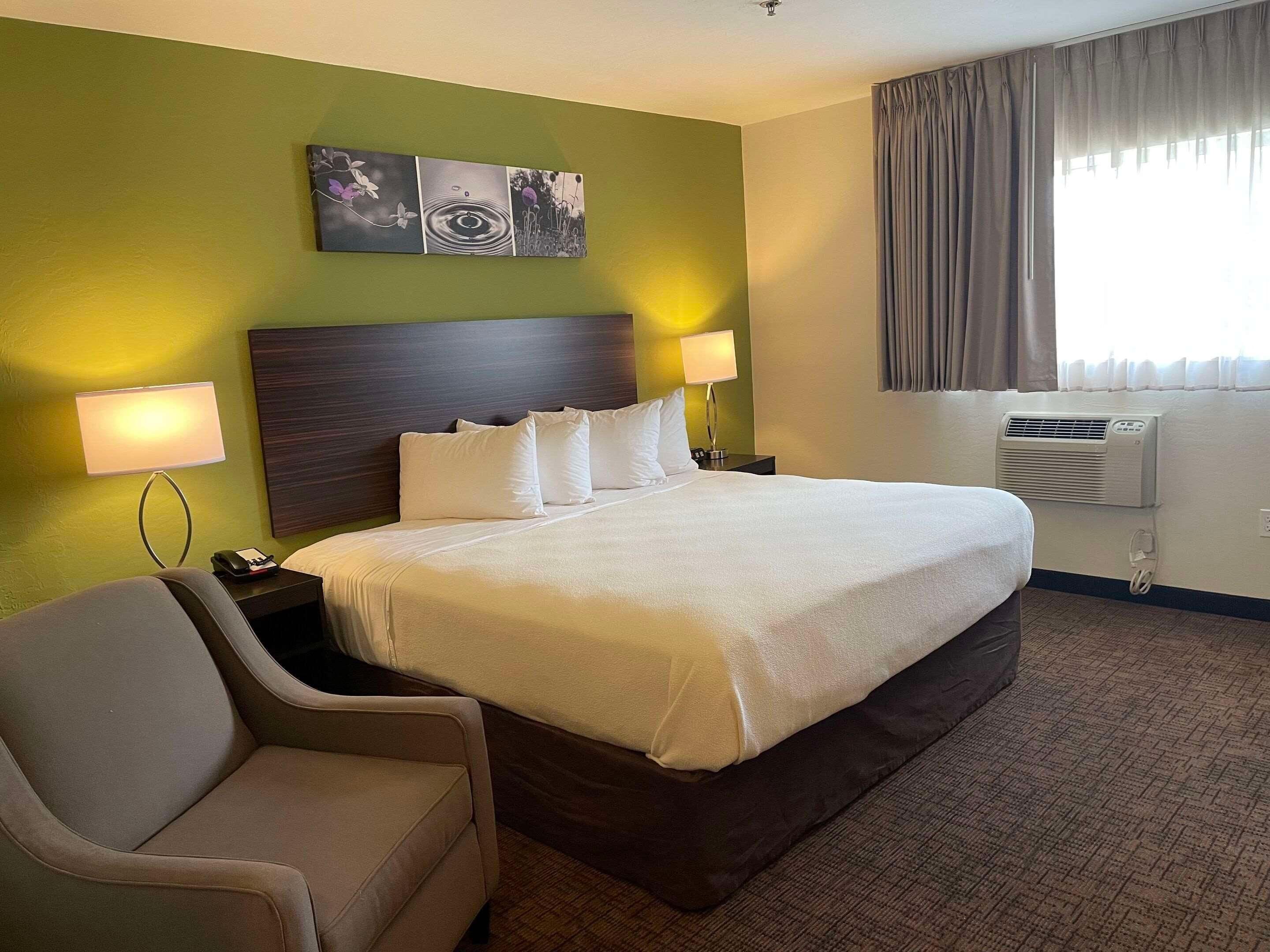 Ein Hotelzimmer mit einem großen Bett, zwei Nachttischlampen, einer Kopfteil, einem Wandbild, einem Fenster mit Vorhängen und einer Klimaanlage.
