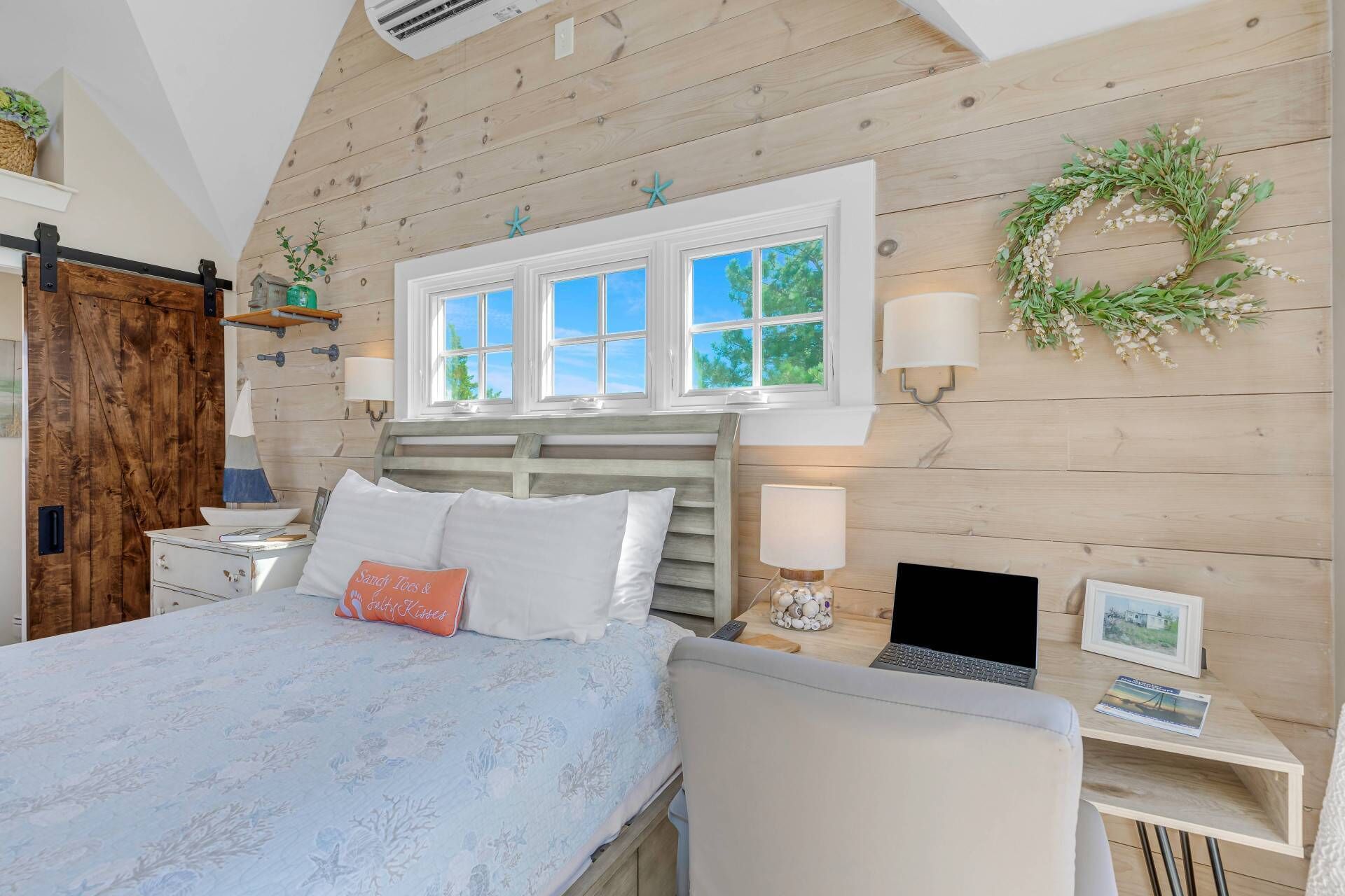 A bedroom with a wooden sloped ceiling, a bed with a floral bedspread, a desk with a laptop, and a window with a view of trees.