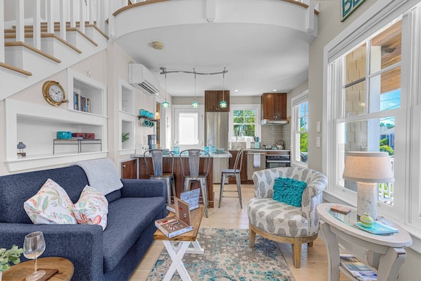 Un salon moderne avec un canapé, un fauteuil à motifs et une table basse. On aperçoit une cuisine à l’arrière-plan, avec un coin repas.