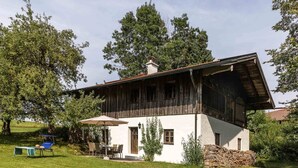 Une maison à deux étages, avec une façade blanche et un étage supérieur en bois foncé. On y trouve une terrasse avec une table et des chaises, ainsi qu’un fauteuil de jardin bleu.