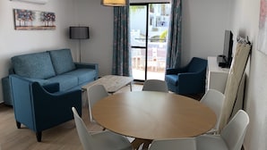 Ett modernt vardagsrum med en blå soffa, ett soffbord i trä och vita stolar. Det finns en platt-TV på ett TV-bord, en lampa och en tavla på väggen.