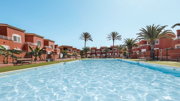 A swimming pool surrounded by red buildings with balconies, palm trees, and a clear sky. - Accommodation "Tucanes" with Terrace and Wi-Fi (Corralejo)