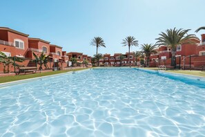A swimming pool surrounded by red buildings with balconies, palm trees, and a clear sky.