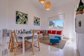 A dining area with wooden chairs and a table, a sofa with red cushions, and two paintings on the wall.