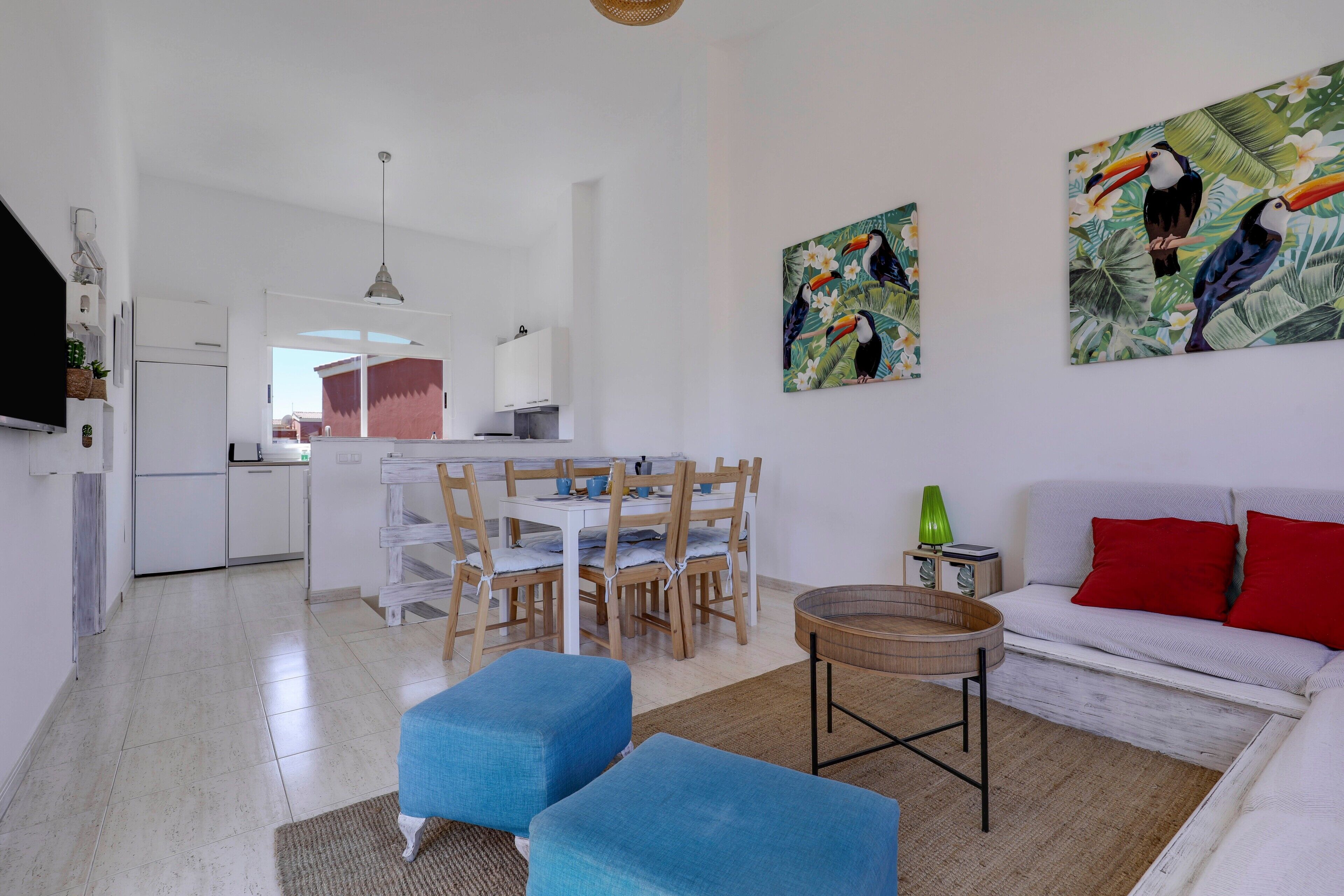 A modern living room with a dining area, white walls, and tropical-themed artwork.