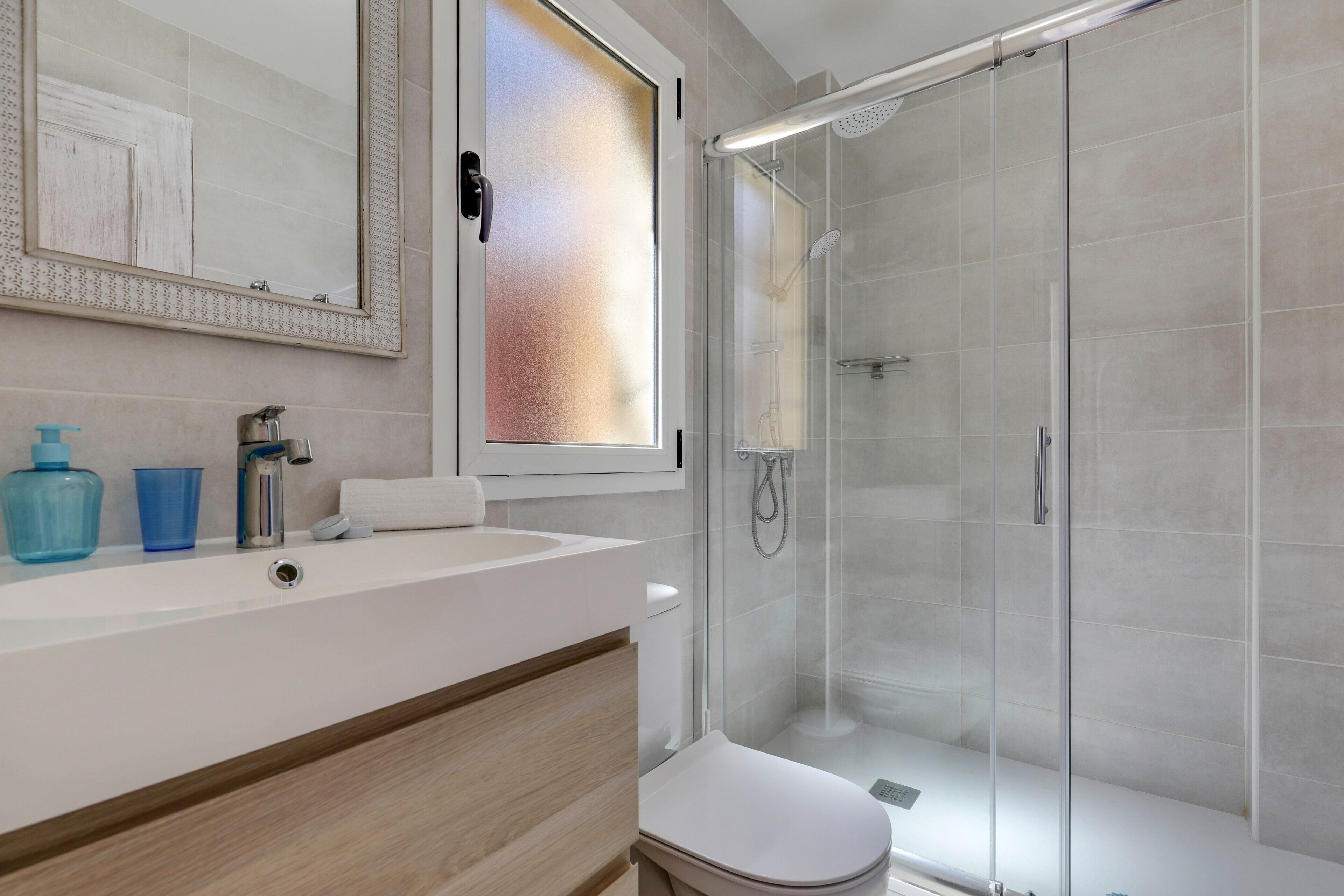 A modern bathroom with a glass shower enclosure, a white sink, and a mirror with a frosted window.