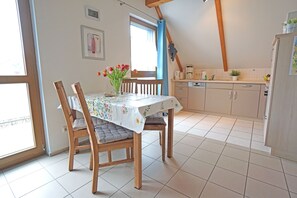 A dining area with a table set for two, wooden chairs, a vase with flowers, and a kitchen in the background.