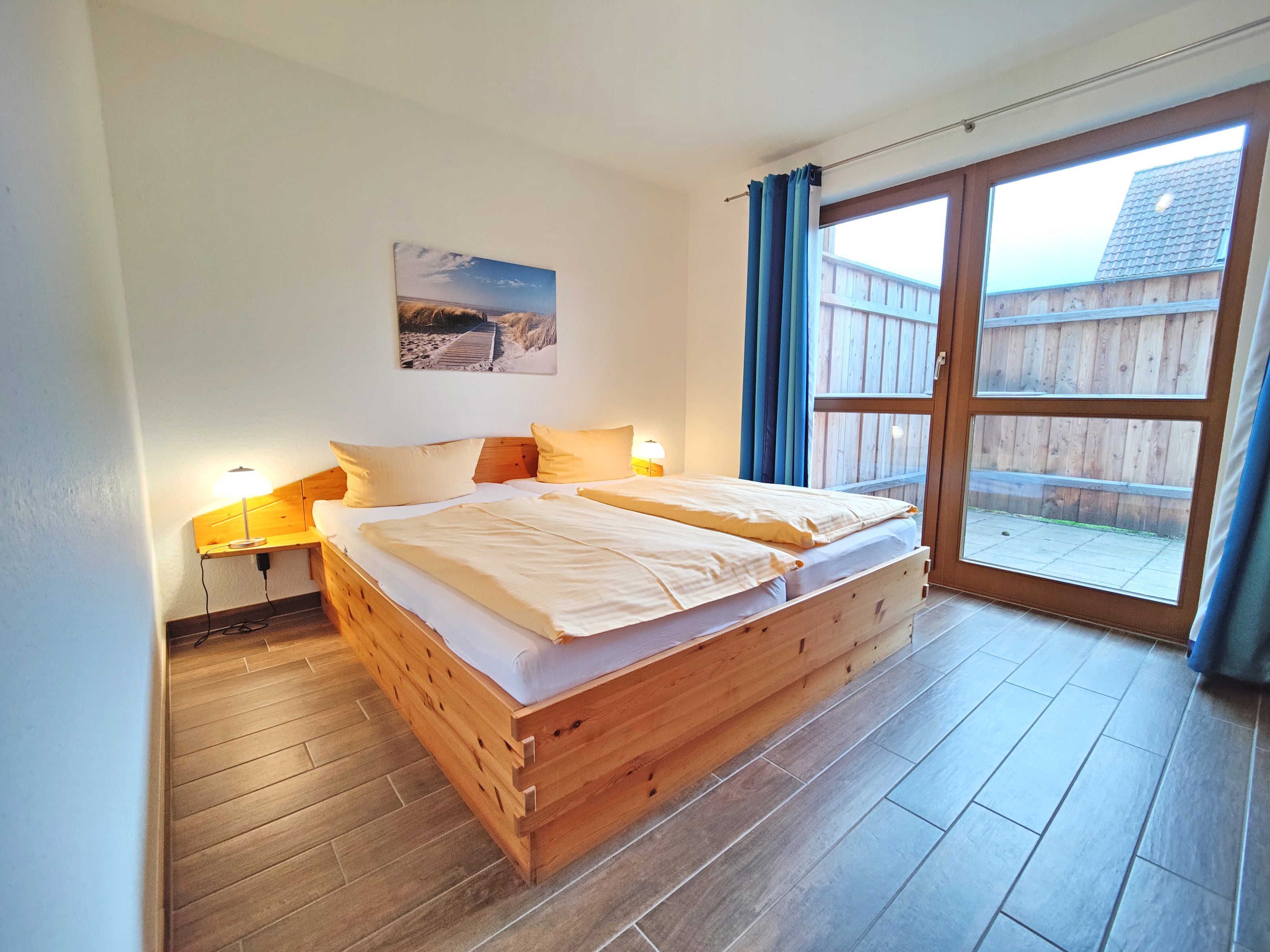 A wooden bed with two pillows, a nightstand with a lamp, a window with curtains, and a picture on the wall.