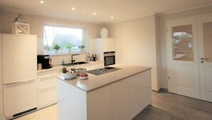 Een moderne keuken met een centraal kookeiland, witte kastjes, een raam met planten en een deur met geëtst glas.