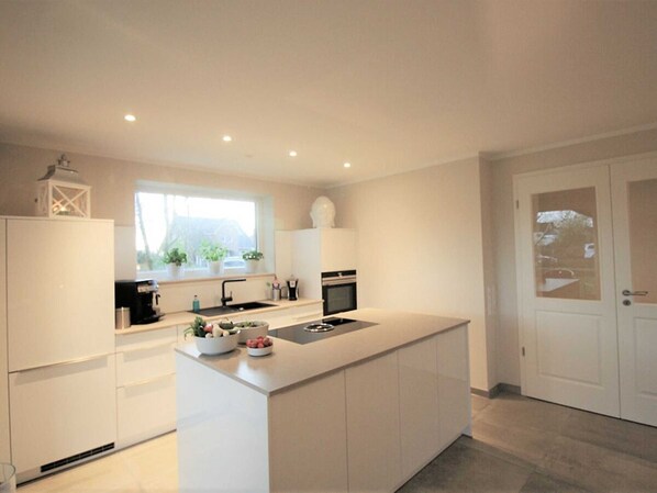 A modern kitchen with a central island, white cabinetry, a window with plants, and a door with a frosted glass panel.