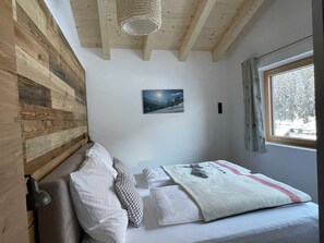 Een slaapkamer met een houten wand, een bed met wit beddengoed, een raam met uitzicht op bomen en een schilderij aan de muur.