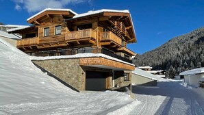 A multi-story wooden chalet with balconies, a stone foundation, and a ski slope in the background. - Apartment Edelweiss - TUX MOUNTAIN apart (Juns)