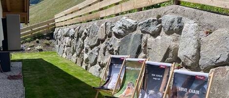 Outdoor seating with posters, a stone wall, and a grassy area with a mountain backdrop.