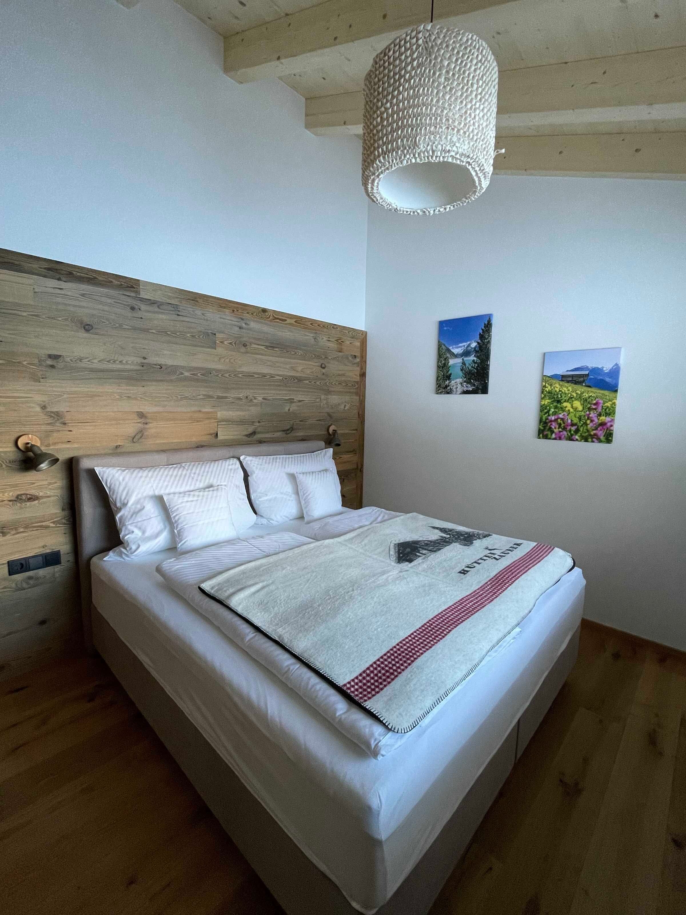 A bedroom with a wooden headboard, a bed with white bedding, a hanging light fixture, and two landscape paintings on the wall.