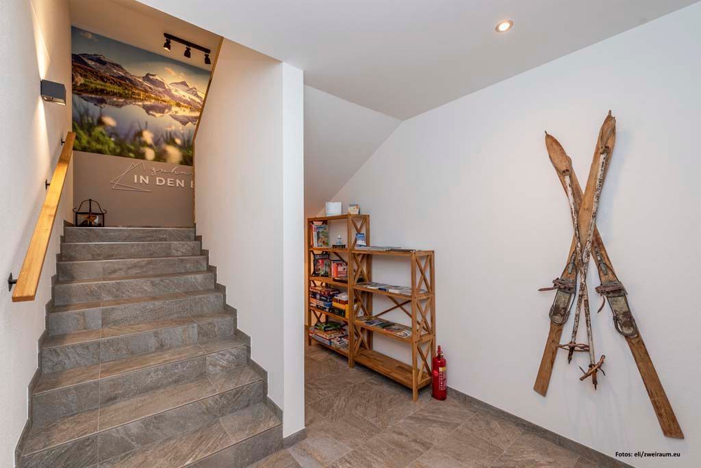 Una escalera conduce a una habitación con un cartel de paisaje montañoso, una estantería de madera y esquíes colgados en la pared.