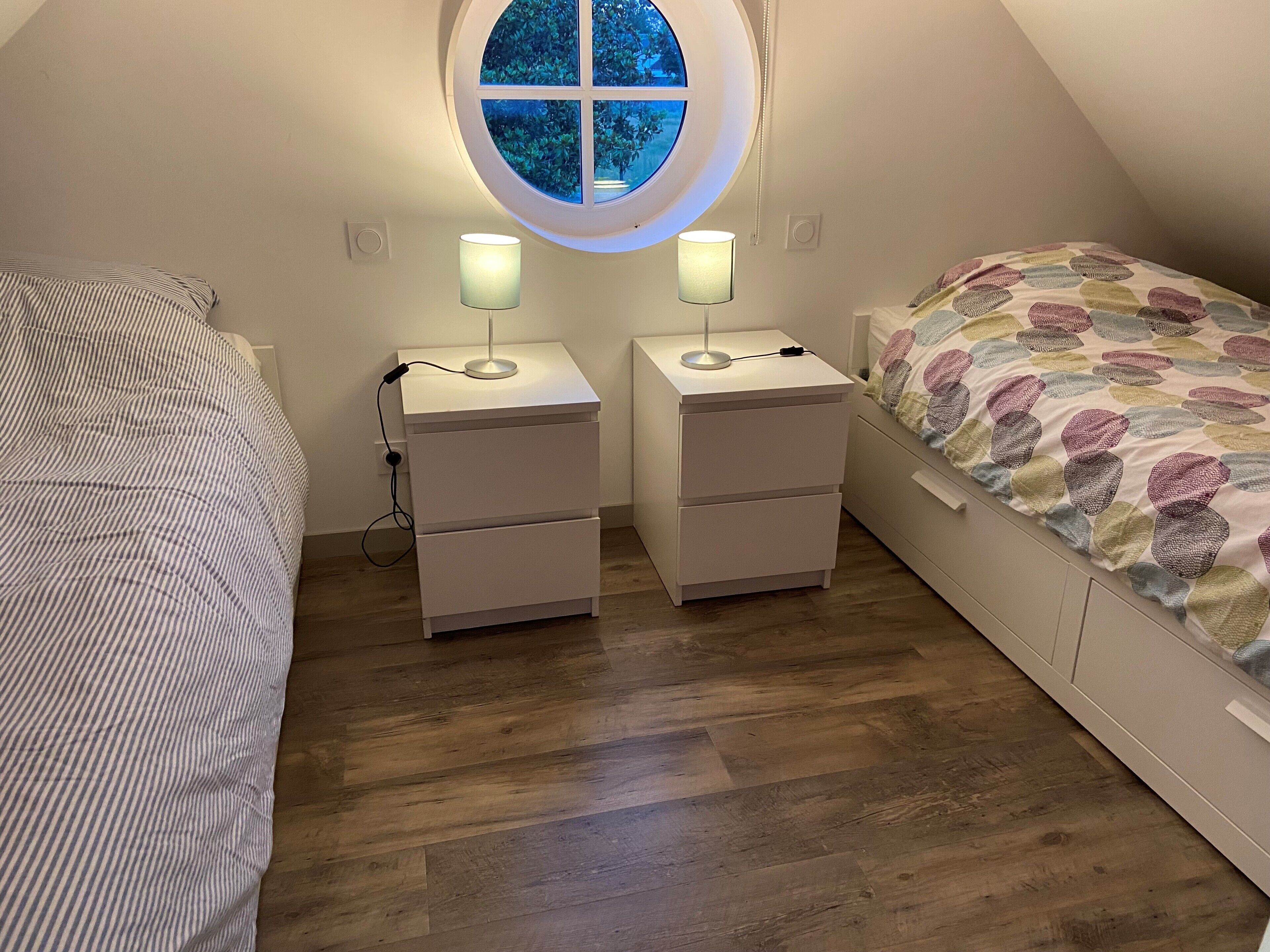 Une chambre avec un plafond en pente, deux tables de chevet avec des lampes, un lit recouvert d’une couverture colorée et une fenêtre donnant sur la forêt.