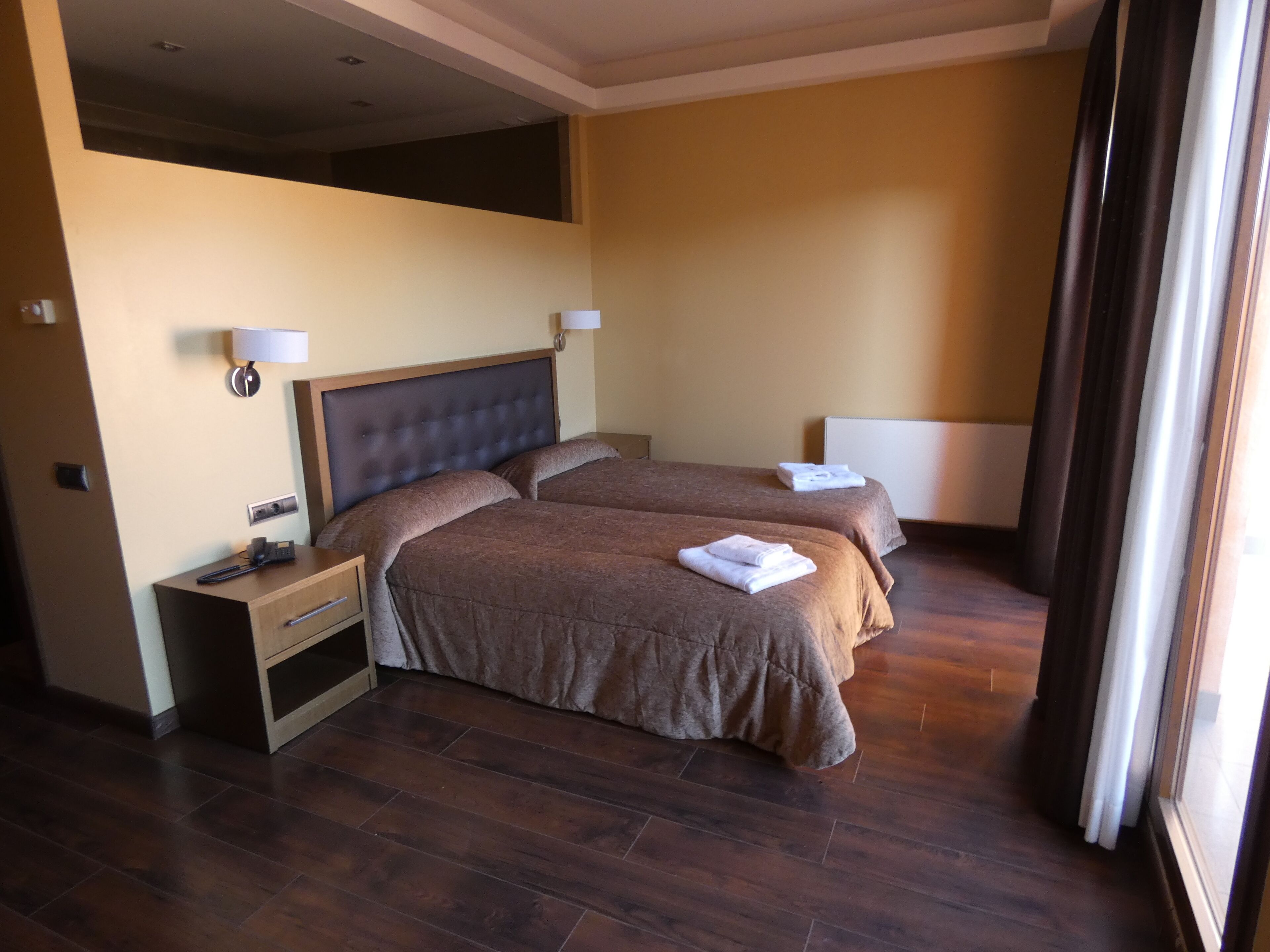 Habitación de hotel con dos camas, una mesita de noche, una lámpara y un ventanal con cortinas.