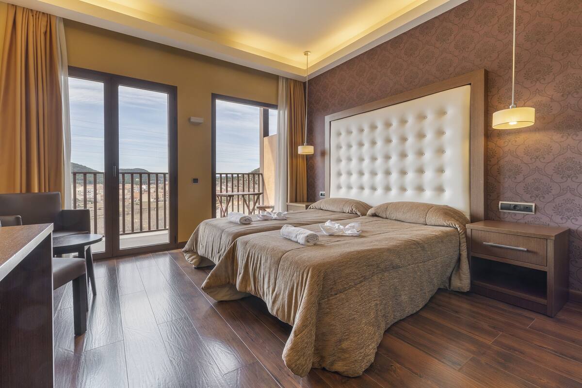 A hotel room with two beds, a desk, and a balcony with a view.
