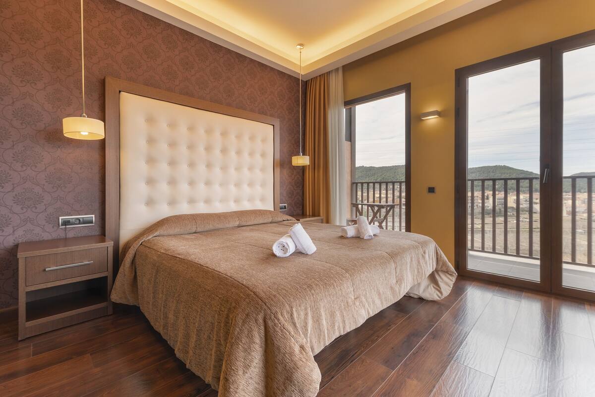 A spacious bedroom with a large bed, a nightstand, a dresser, and a balcony with a view of the hills.