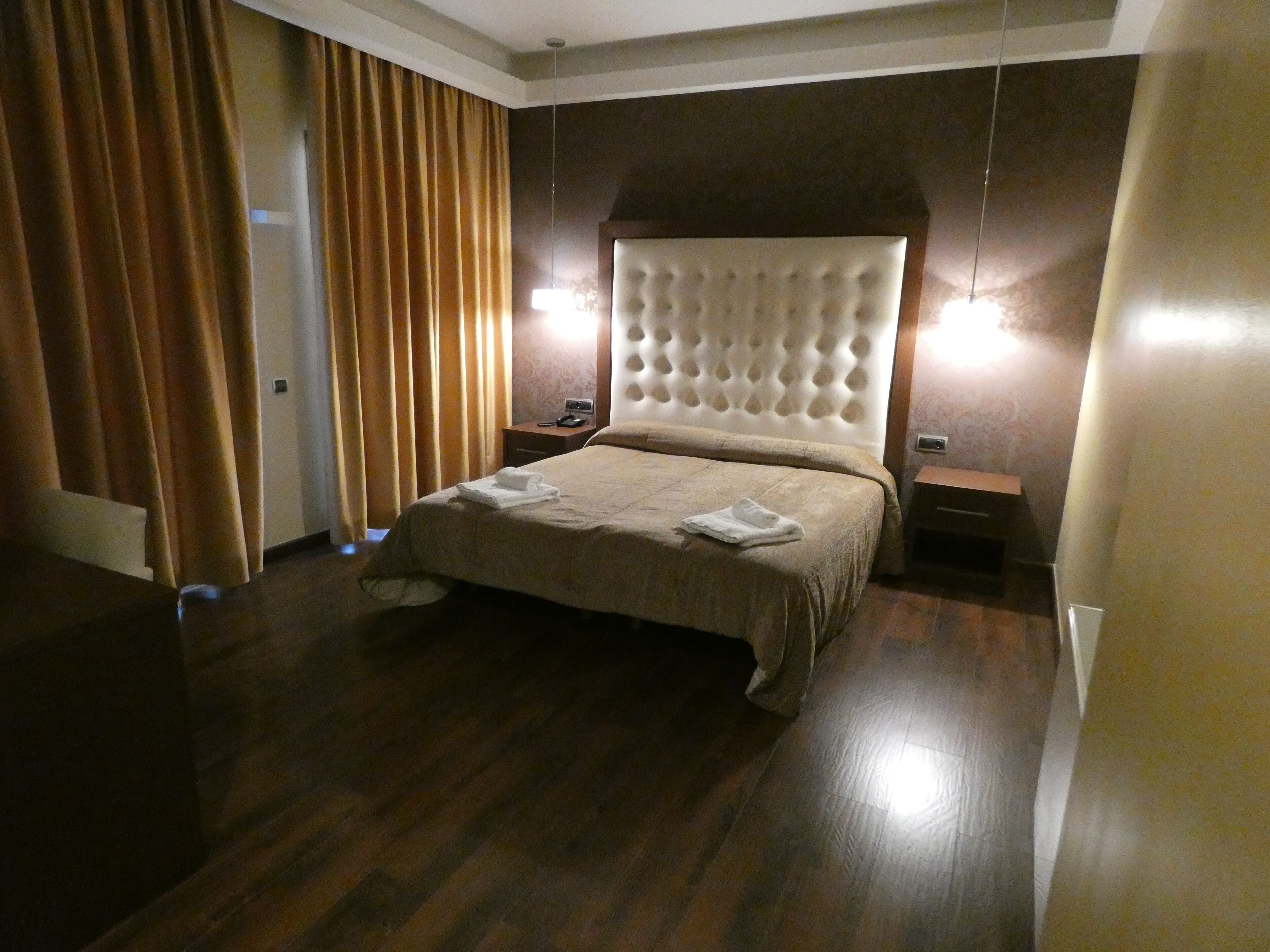 A hotel room with a large bed, wooden flooring, a headboard with a tufted design, and two bedside tables with lamps.