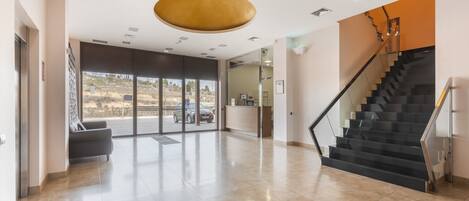 A spacious lobby with a reception desk, a staircase, and a large window offering a view of the outdoors.