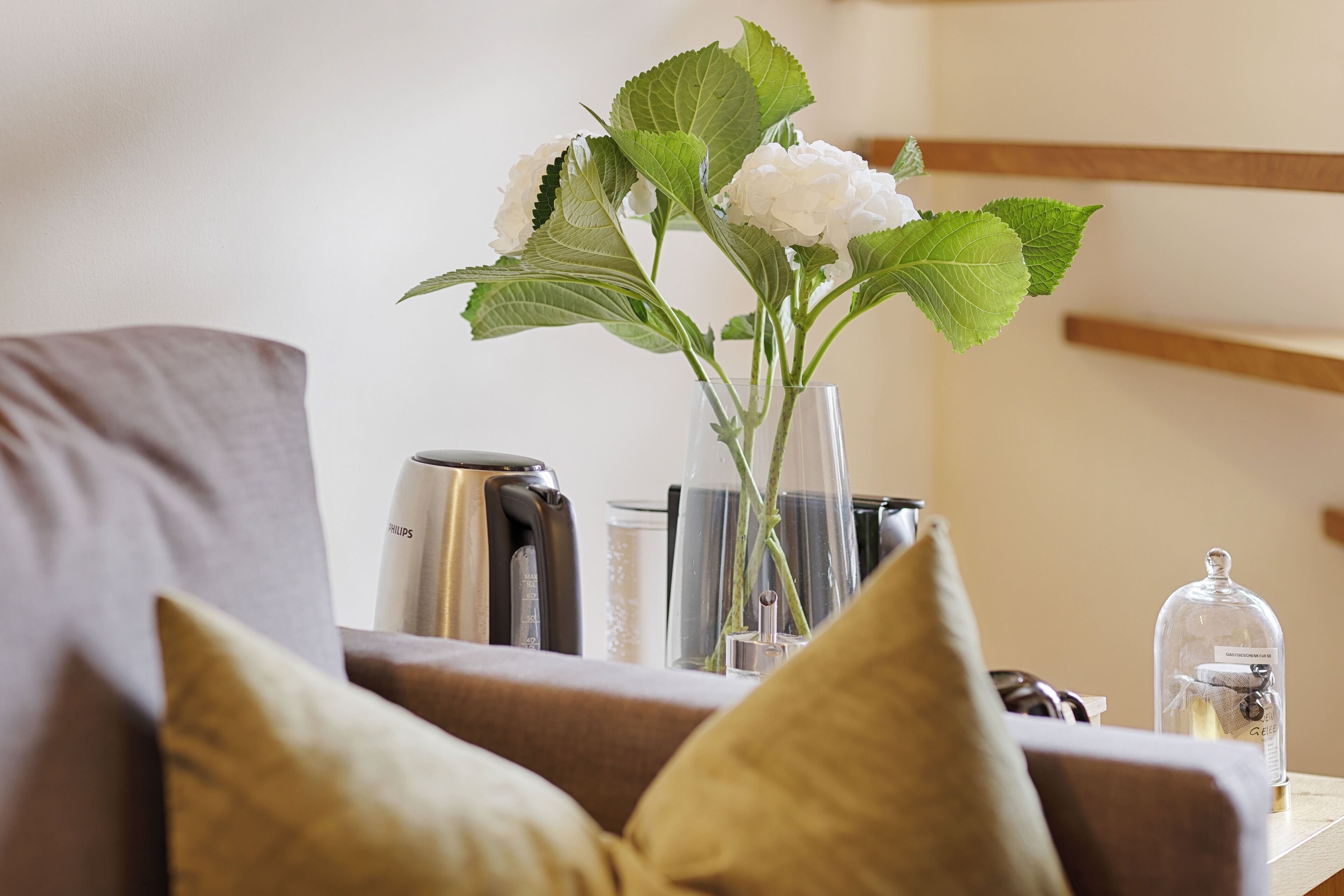 A modern living room with a sofa, a glass vase with white flowers, a coffee maker, and a glass jar on a wooden table.