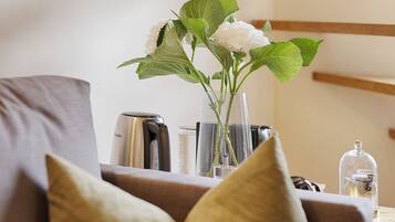 A modern living room with a sofa, a glass vase with white flowers, a coffee maker, and a glass jar on a wooden table.