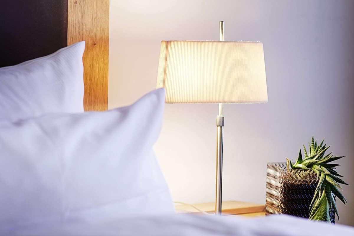 Cama bem arrumada com roupa de cama branca, abajur e uma pequena planta em mesa de cabeceira de madeira.