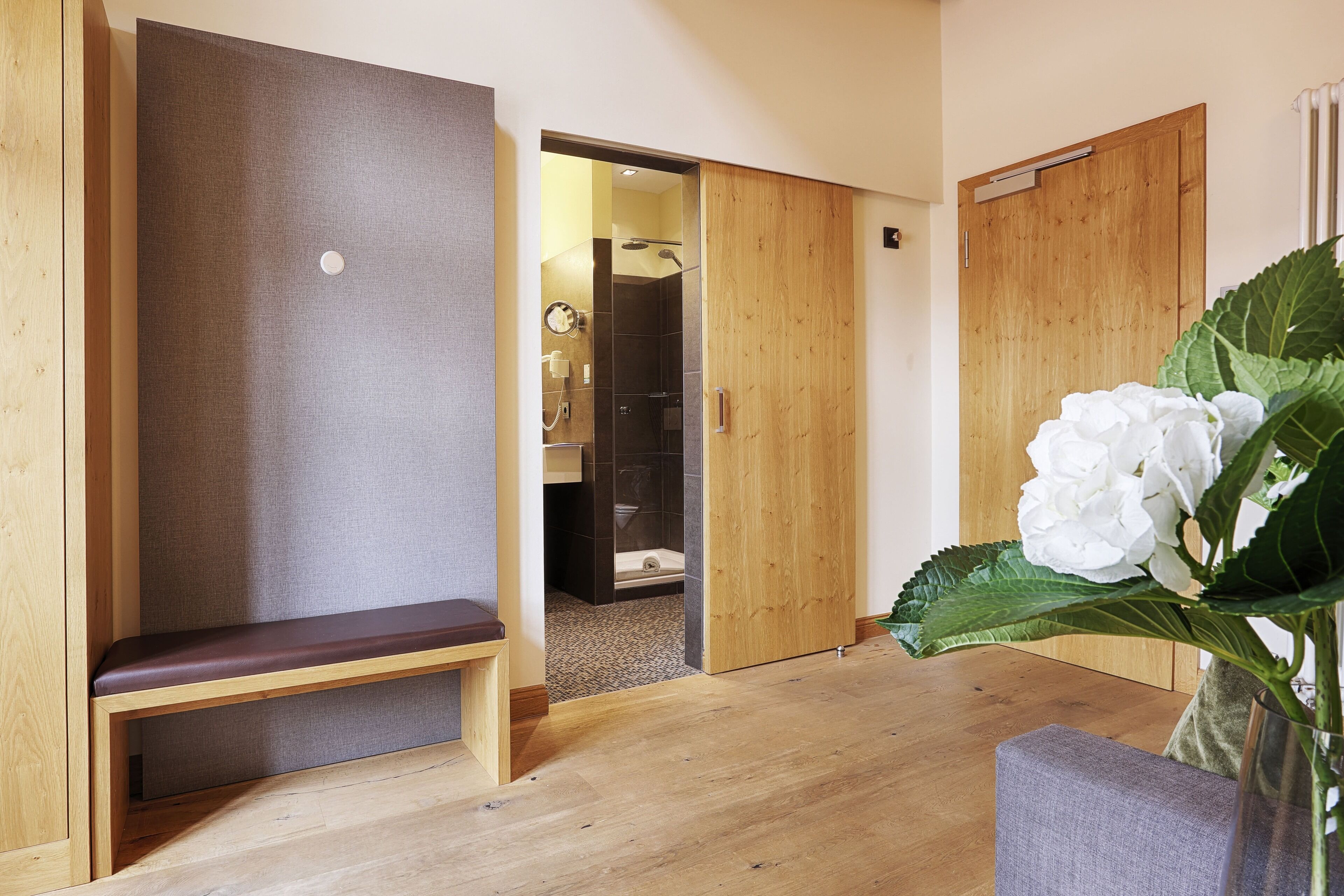 A wooden bench, a wooden door, and a white flower in a room with a wooden floor.