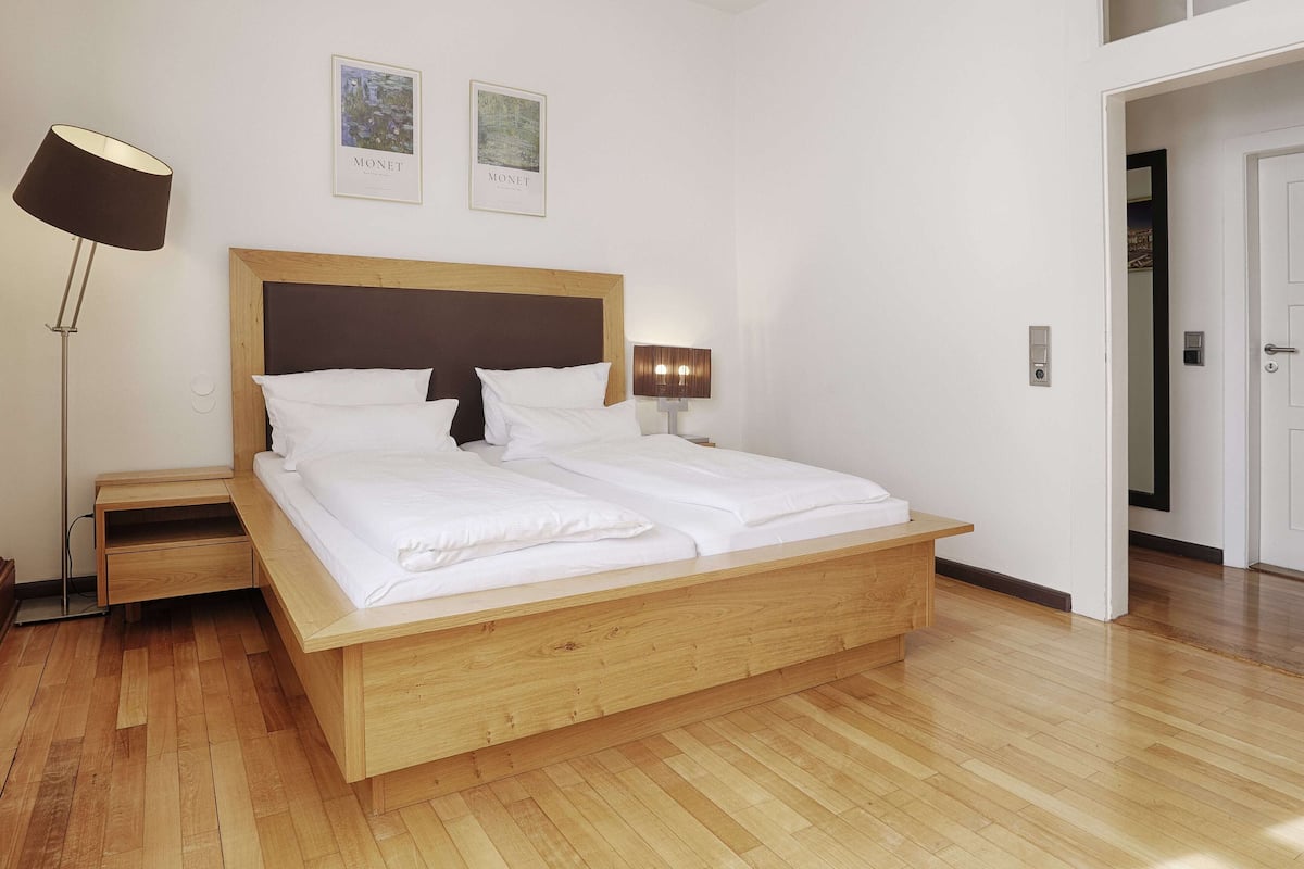 Quarto moderno com cama de madeira, mesas de cabeceira, abajur e duas molduras na parede.