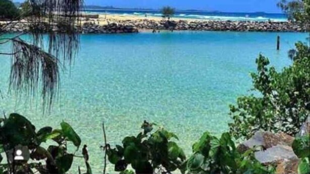 A beach with clear turquoise water, a rocky shoreline, and lush greenery in the foreground. - The Family Beach House (Casuarina)